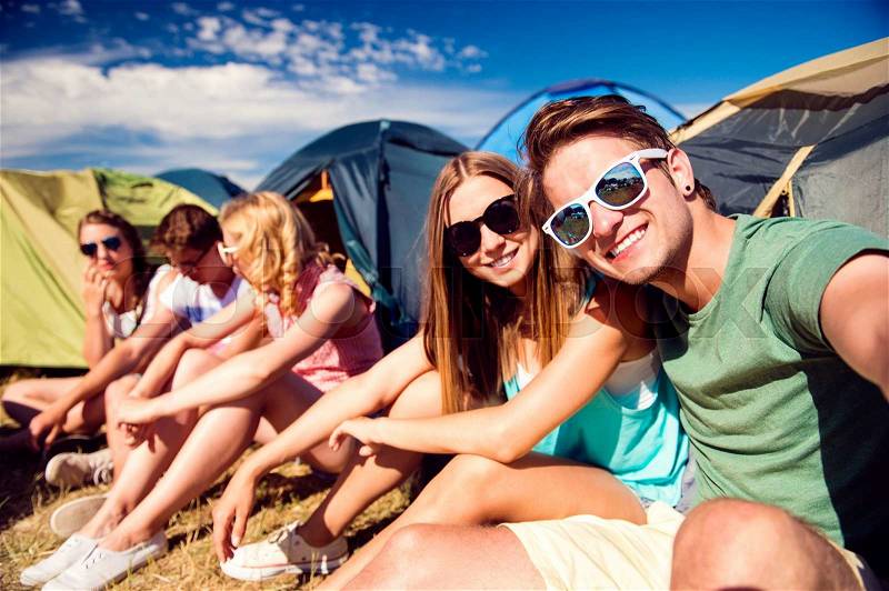 Group of teenage boys and girls at summer music festival, sitting on the ground in front of tents, taking selfie, stock photo