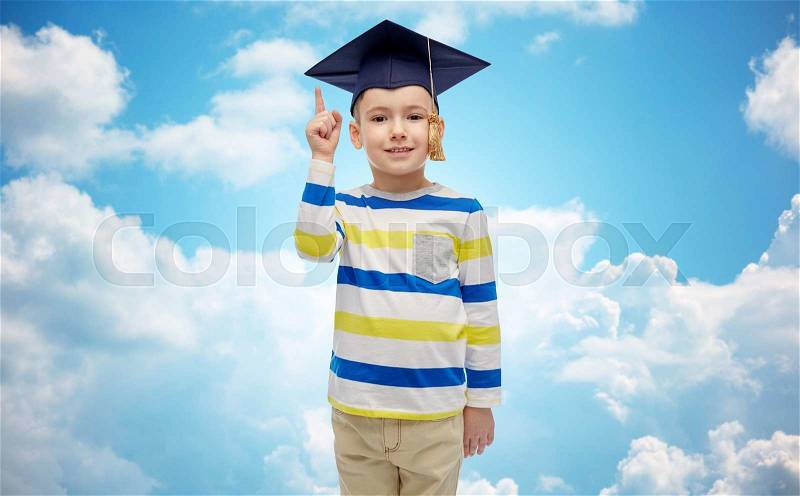 Childhood, school, education, learning and people concept - happy boy in bachelor hat or mortarboard pointing finger up over blue sky and clouds background, stock photo