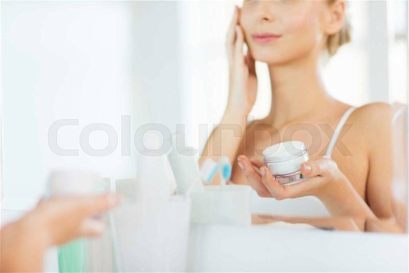 Beauty, skin care and people concept - close up of smiling young woman applying cream to face mirror reflection at home bathroom, stock photo