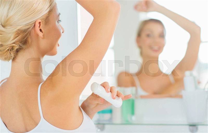 Beauty, hygiene, morning and people concept - close up of smiling young woman applying antiperspirant or stick deodorant and looking to mirror at home bathroom, stock photo
