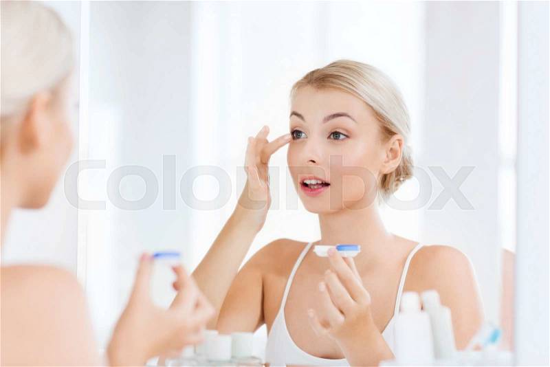 Beauty, vision, eyesight, ophthalmology and people concept - young woman putting on contact lenses at mirror in home bathroom, stock photo