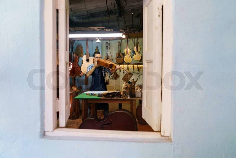 Lute maker shop and classic music instruments: young adult artisan fixing old classic guitar. The man looks carefully at bridge and arm to check the wood curvature. Full length, stock photo