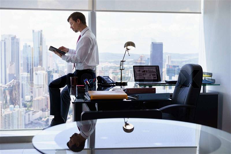 Adult businessman sitting on desk in modern office and reading news on tablet pc. The man works in a skyscraper with a view of the city from the large window, stock photo