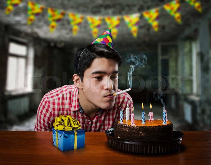 Sad birthday boy smoking in an abandoned room with cake and present, stock photo