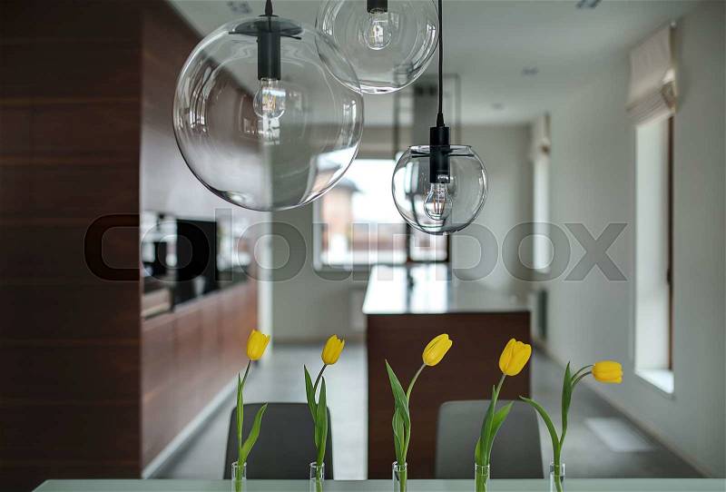 Five yellow tulips in glass vases on the table and three glass round lamps over them. They are on the soft background of brown kitchen with light walls. Horizontal, stock photo