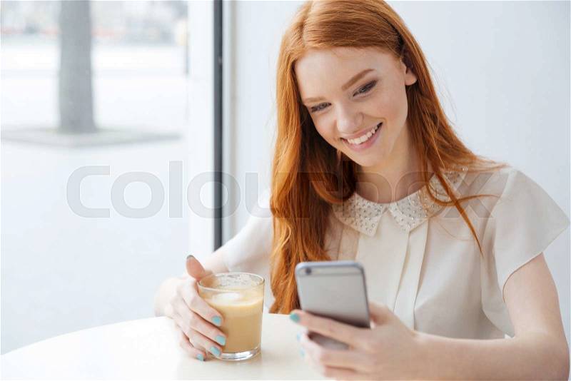Amiling pretty redhead young woman sitting in cafe and using cell phone , stock photo