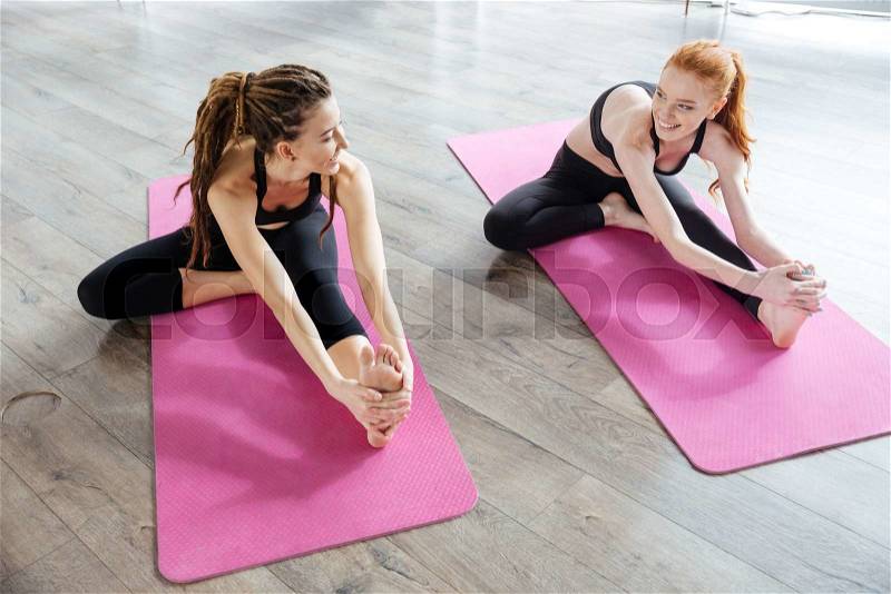 Two happy cute young women sitting and stretching in yoga center, stock photo