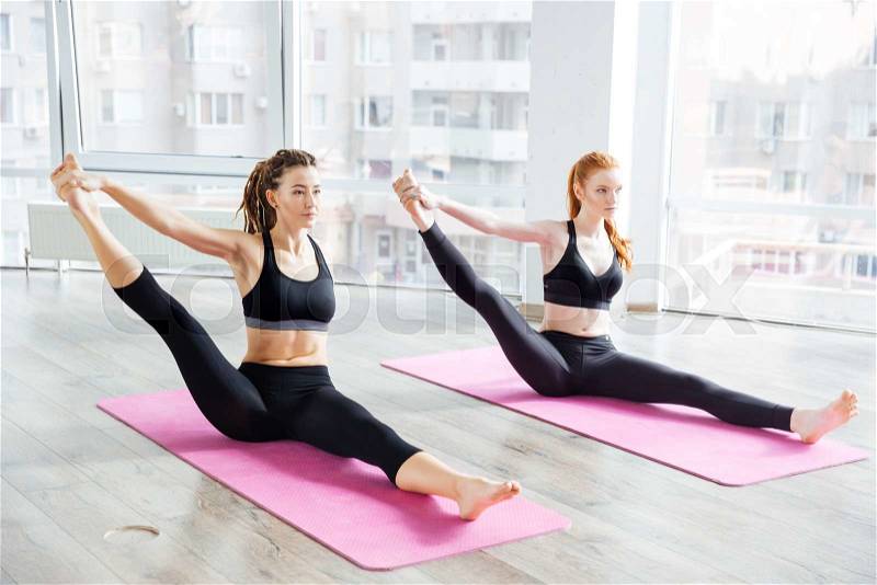 Two attractive young women stretching legs and practicing yoga in studio, stock photo