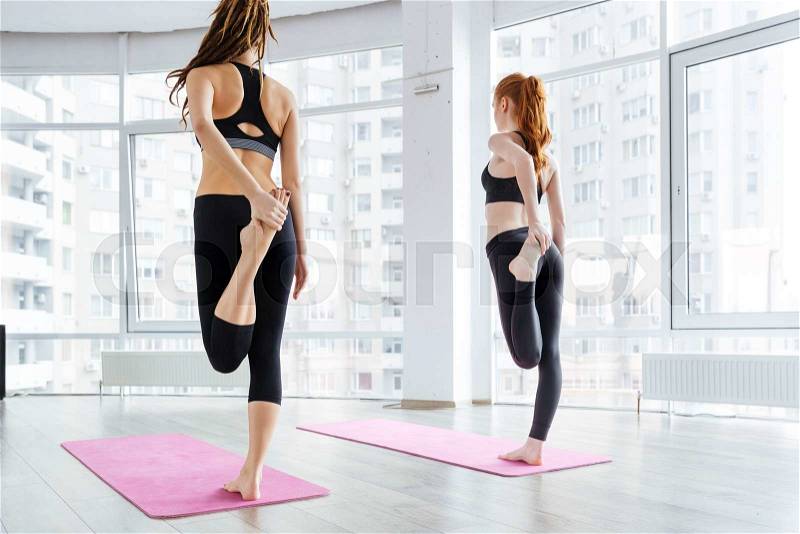 Back view of two attractive young women standing and stretching legs on pink yoga mat, stock photo