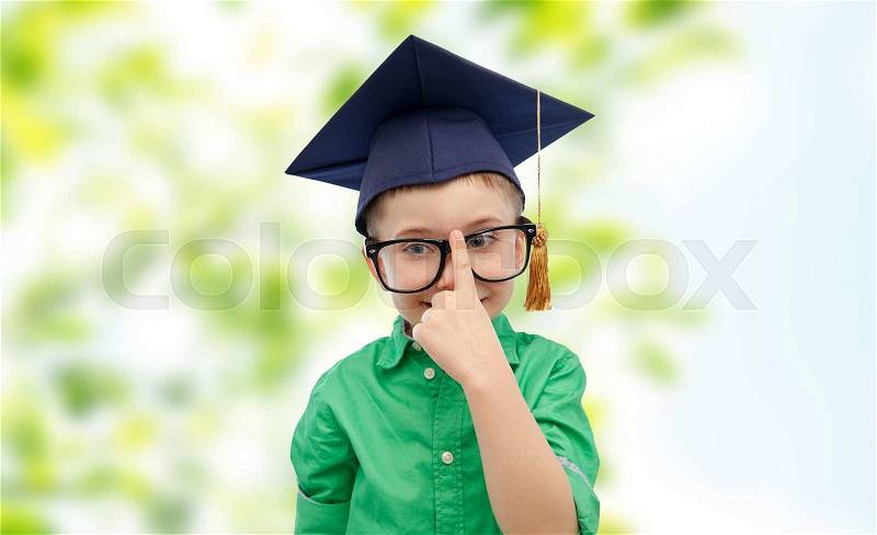 Childhood, school, education, knowledge and people concept - happy boy in bachelor hat or mortarboard and eyeglasses over green natural background, stock photo