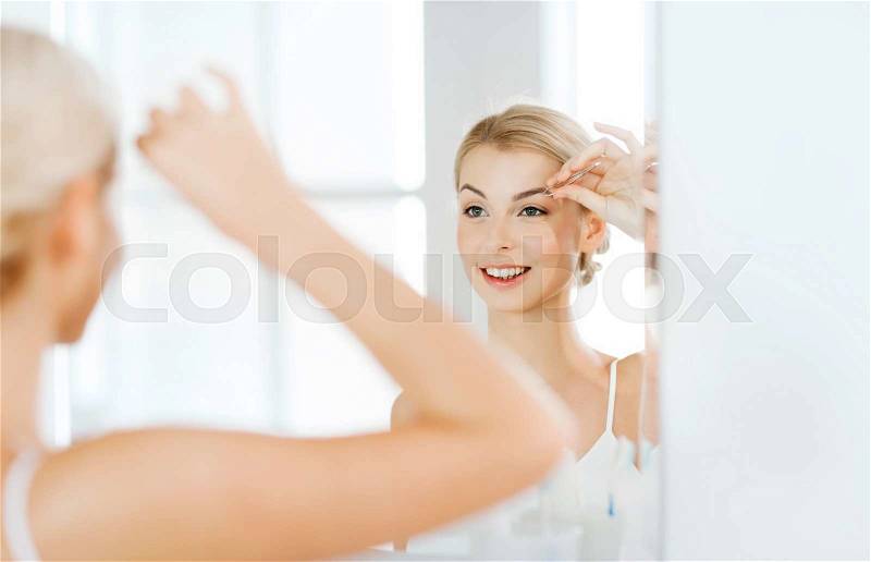 Beauty and people concept - smiling young woman with tweezers tweezing eyebrow and looking to mirror at home bathroom, stock photo