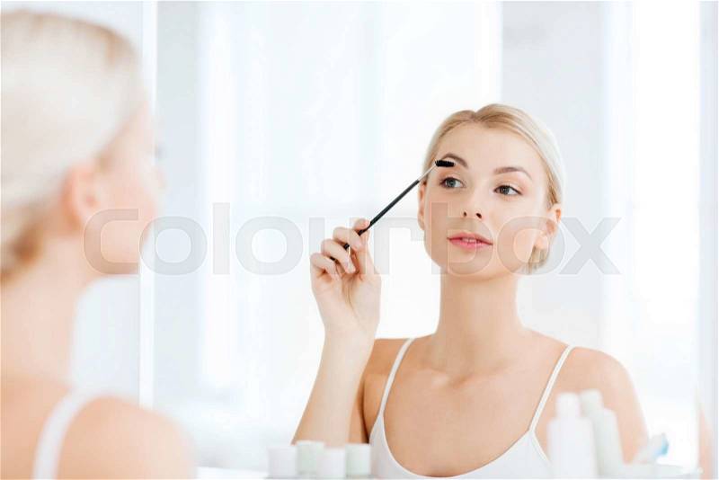 Beauty, make up, morning and people concept - young woman brushing eyebrow with brush and looking to mirror at home bathroom, stock photo