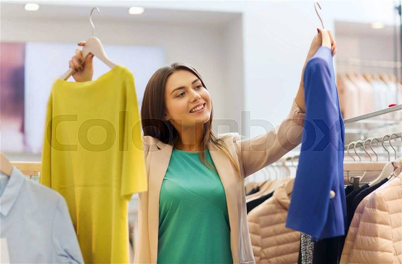 Sale, clothes , shopping, fashion and people concept - happy young woman choosing between shirt and jacket at clothing store, stock photo