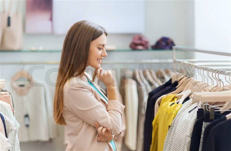 Sale, shopping, fashion, style and people concept - happy young woman choosing clothes in mall or clothing store, stock photo