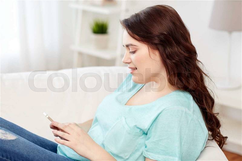 People, technology, communication and leisure concept - happy young plus size woman sitting on sofa with smartphone at home, stock photo