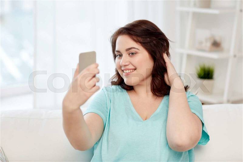 People, technology, communication and leisure concept - happy young plus size woman taking selfie with smartphone at home, stock photo
