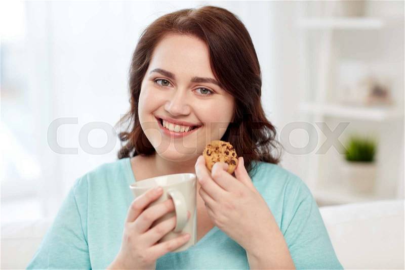 People, junk food, unhealthy eating and leisure concept - happy plus size young woman with cup of tea with cookie at home, stock photo