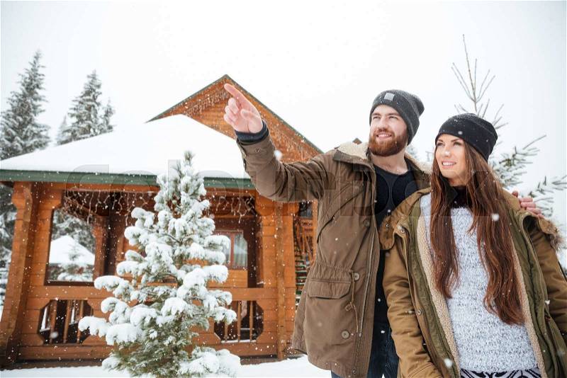 Hamdsome smiling bearded young man stading with his girlfriend and pointing away near log cabin, stock photo