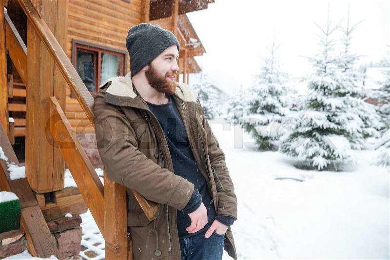 Thoughtful handsome young man with beard standing near log cabine in winter , stock photo