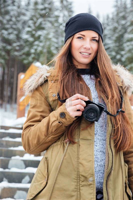 Portrait of happy beautiful woman with photo camera standing on stairs in winter forest, stock photo