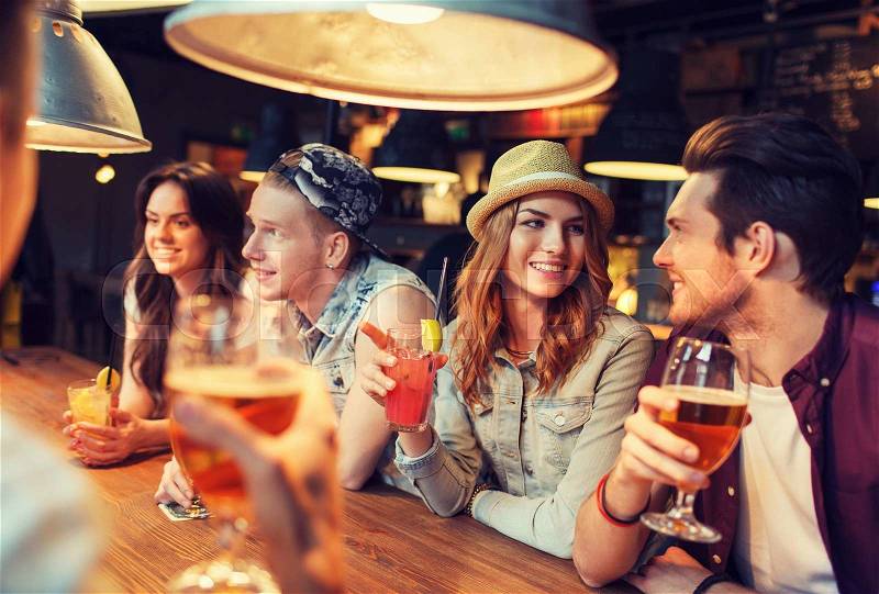 People, leisure, friendship and communication concept - group of happy smiling friends drinking beer and cocktails talking at bar or pub, stock photo