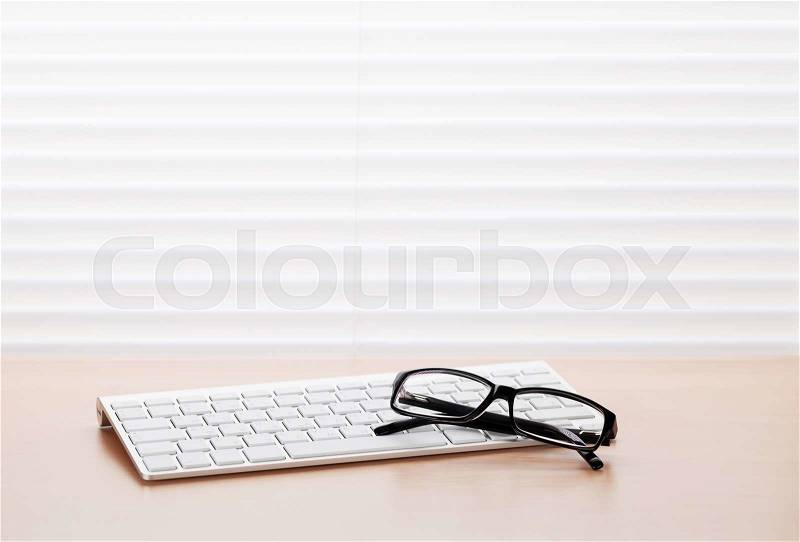 Office workplace with pc keyboard and glasses on wooden desk table in front of window with blinds. View with copy space, stock photo