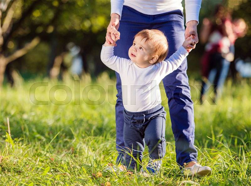 Unrecognizable mother holding hands of her little son making first steps outside in green sunny nature, stock photo