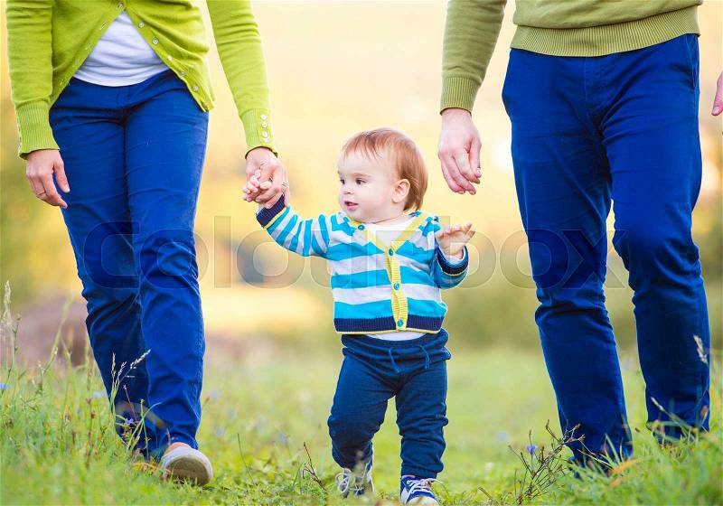 Unrecognizable mother and father holding hands of their little son making first steps outside in green sunny nature, stock photo
