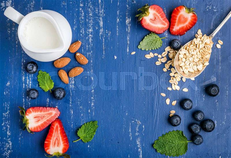 Healthy breakfast ingredients food frame. Oatmeal in spoon, milk in creamer, berries, almond and mint on painted blue wooden background, top view, copy space, stock photo