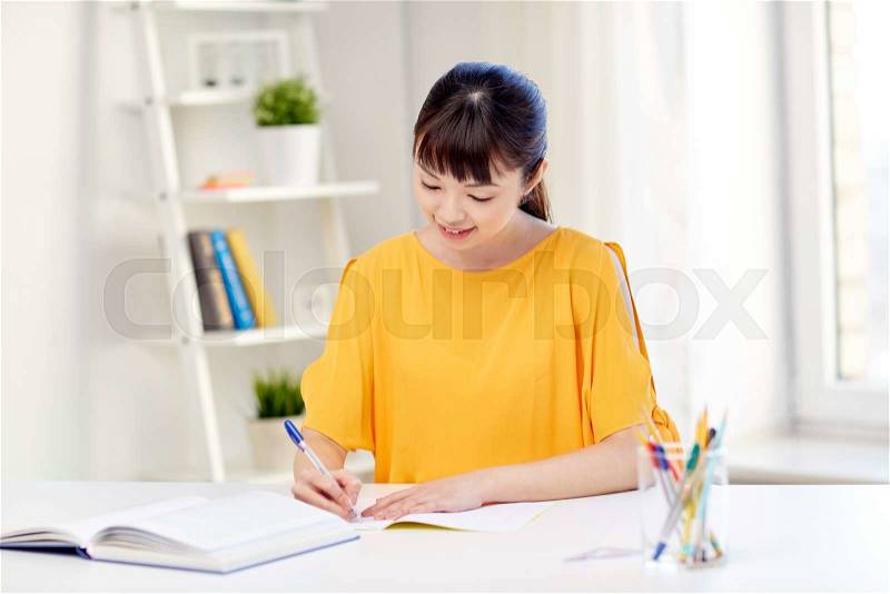 People, education, high school and learning concept - happy asian young woman student with book and notepad writing at home, stock photo
