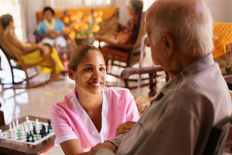 Old people in geriatric hospice: young attractive hispanic woman working as nurse takes care of a senior man on wheelchair. She talks with him then goes away to help other patients, stock photo