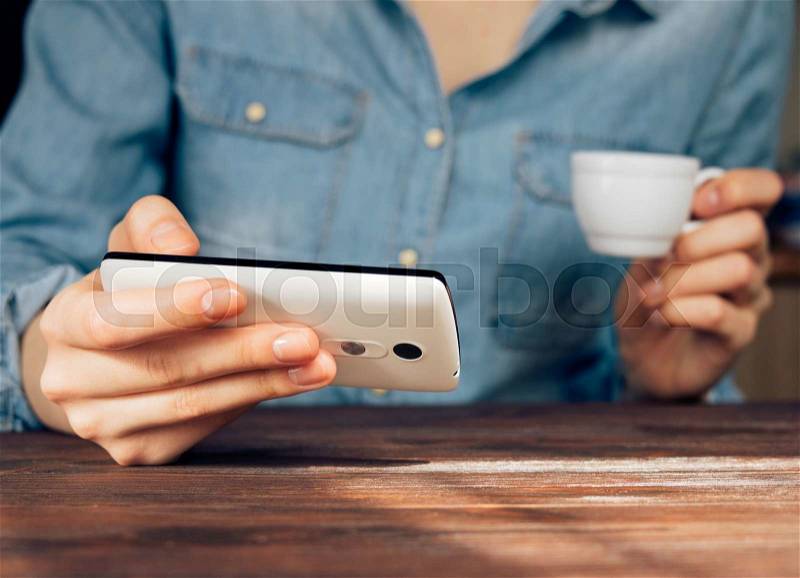 Woman in a denim shirt reading news on mobile phone and drinking coffee, stock photo