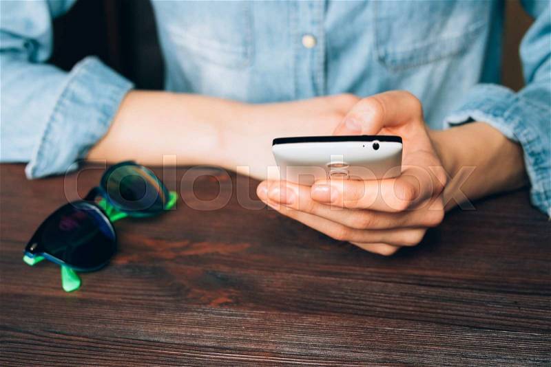 Woman in a denim shirt holds a mobile phone and sitting at a wooden table on which lie sunglasses, stock photo