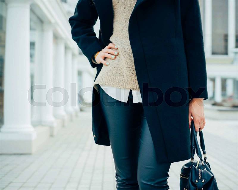 Woman in an elegant coat holding a handbag in one hand, the other hand on her waist. In the background details of ancient architecture, stock photo