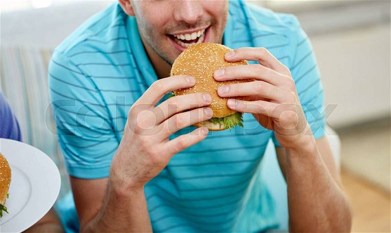 Fast food, unhealthy eating, people and junk-food - close up of happy man eating hamburger at home, stock photo