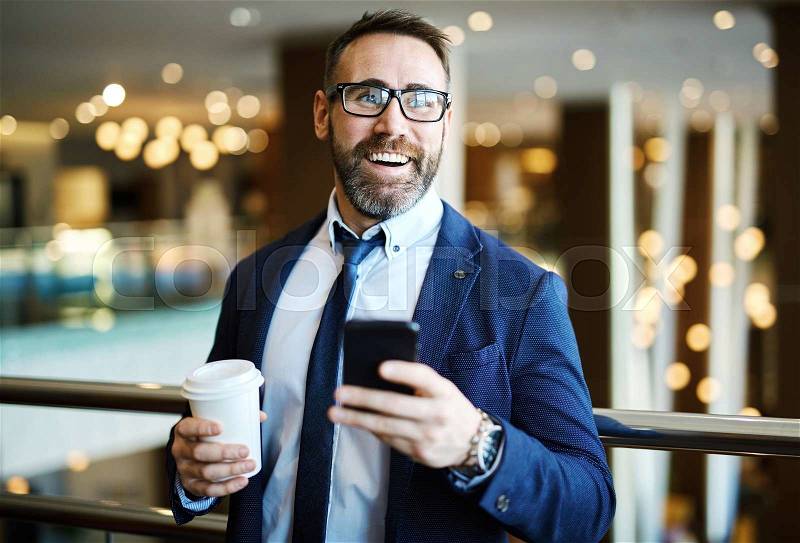 Happy businessman holding glass with coffee and cellphone, stock photo