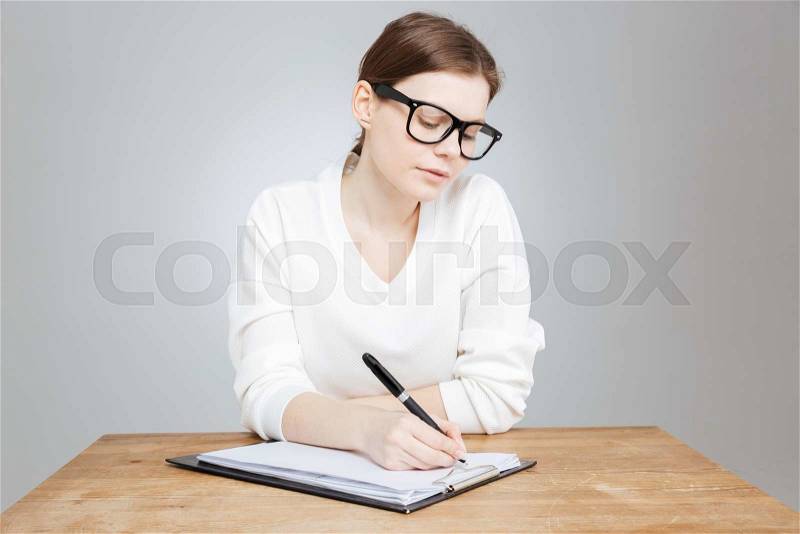 Thoughtful pretty teenage girl in glasses sitting and writing on clipboard over gray, stock photo