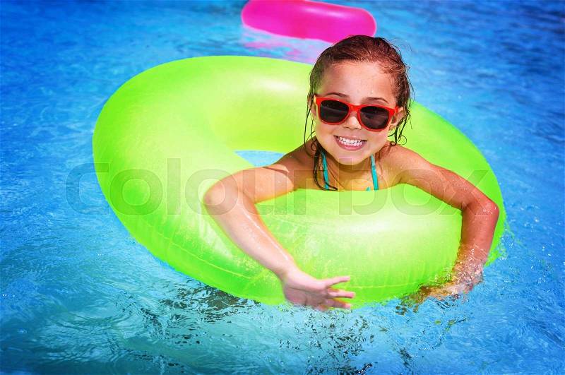 Portrait of cute happy little girl having fun in swimming pool, floating in blue refreshing water with big green rubber ring, active summer vacation on the beach, stock photo