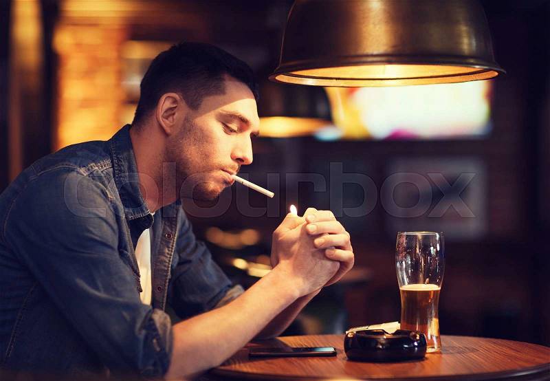 People, smoking and bad habits concept - man drinking beer and lighting cigarette at bar or pub, stock photo