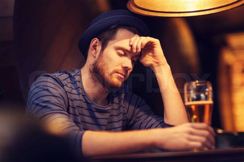 People, loneliness, alcohol and lifestyle concept - unhappy single young man in hat drinking beer at bar or pub, stock photo