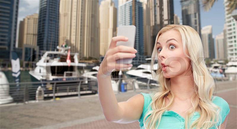Expressions, travel, tourism, technology and people concept - funny young woman or teenage girl taking selfie with smartphone and making fish face over boats in harbor in dubai city background, stock photo