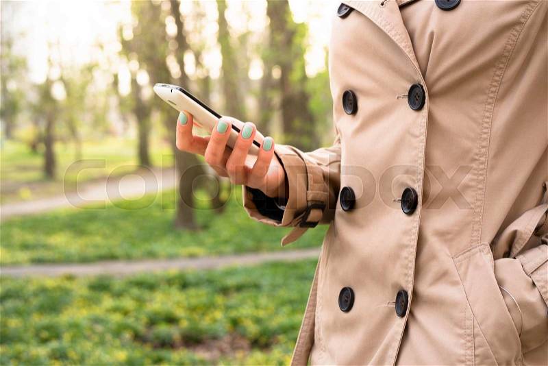 Woman in beige coat walks in the park and using a mobile phone, stock photo