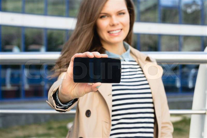 Attractive smiling woman shows the display of the smart phone outdoors. The girl wearing a beige jacket and a striped shirt, stock photo