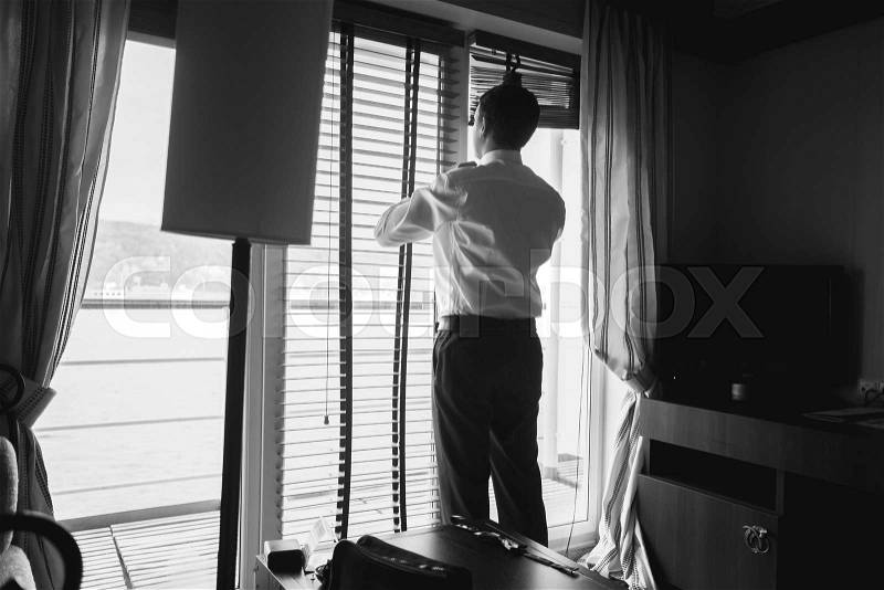 Black and white portrait of stylish businessman looking out of window at hotel room, stock photo