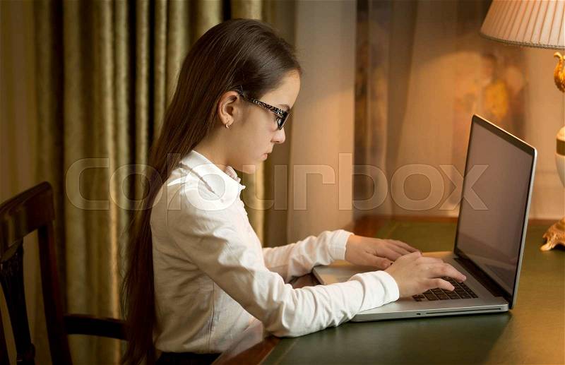Teen girl in school uniform sitting behind table and using laptop at cabinet, stock photo