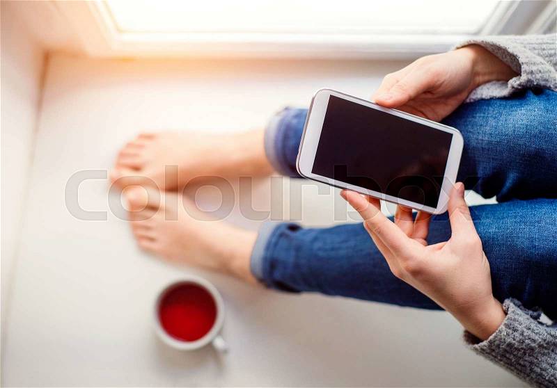 Unrecognizable woman sitting on window sill with smart phone, texting, stock photo