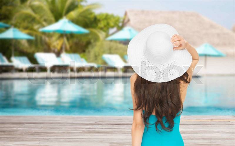 People, summer holidays, travel, tourism and vacation concept - woman in swimsuit and sun hat from back over exotic hotel resort beach with swimming pool and sunbeds background, stock photo