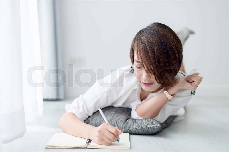 Happy Asian woman lying down on floor and writting at home, stock photo