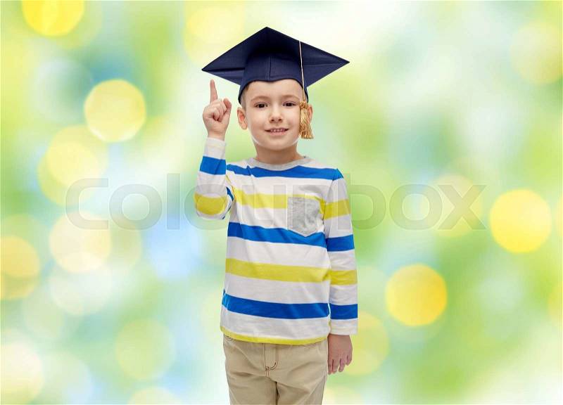 Childhood, school, education, learning and people concept - happy boy in bachelor hat or mortarboard pointing finger up over summer green lights background, stock photo