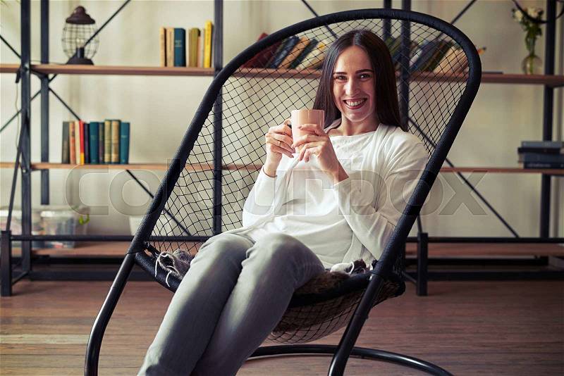 Young caucasian brunette with wide smile sitting in a designer chair with a cup, stock photo